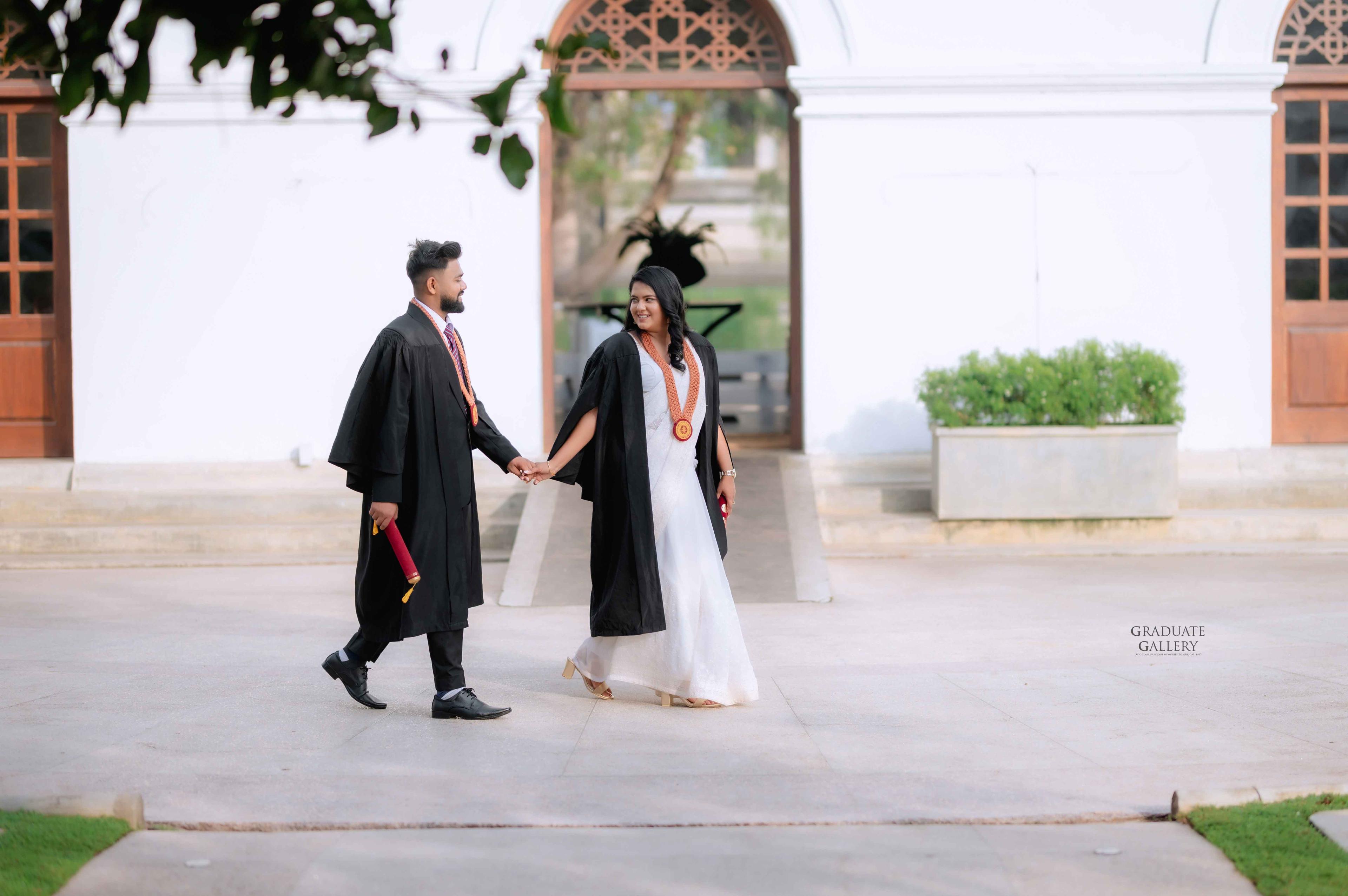 Graduate couple in gowns walking hand in hand past a white colonial building