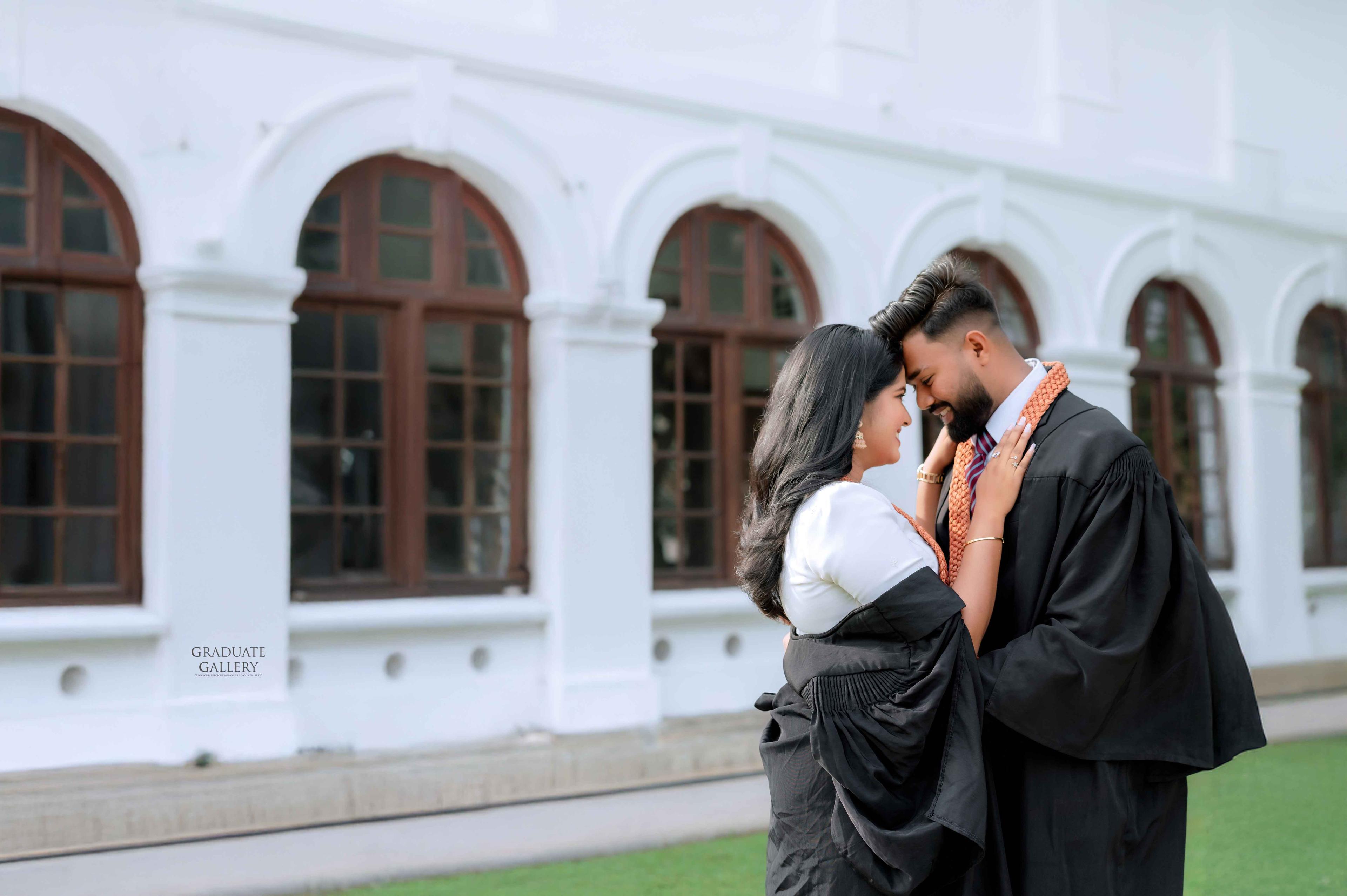 Graduate couple in black gowns sharing a moment beside arched windows