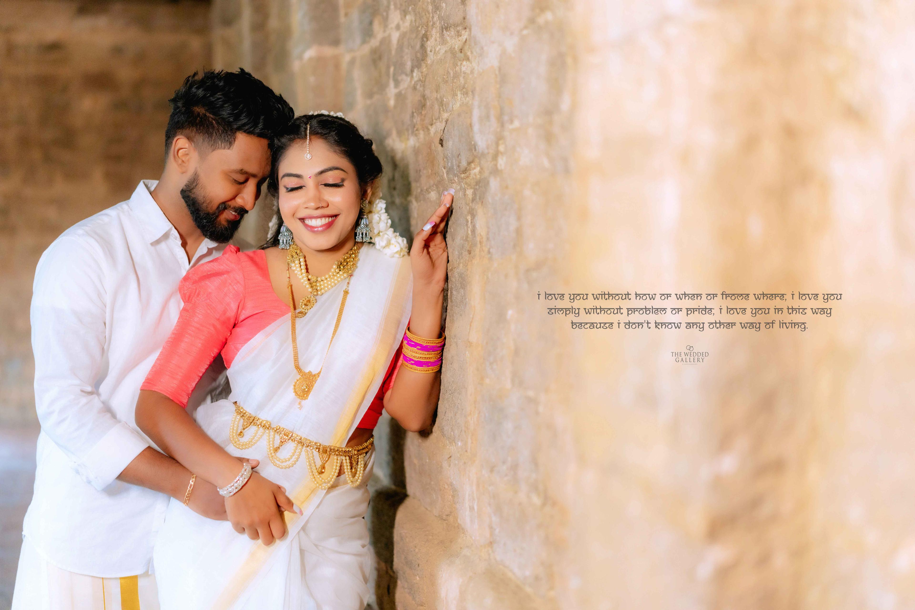 Couple in traditional white and coral attire embracing beside a sandstone pillar